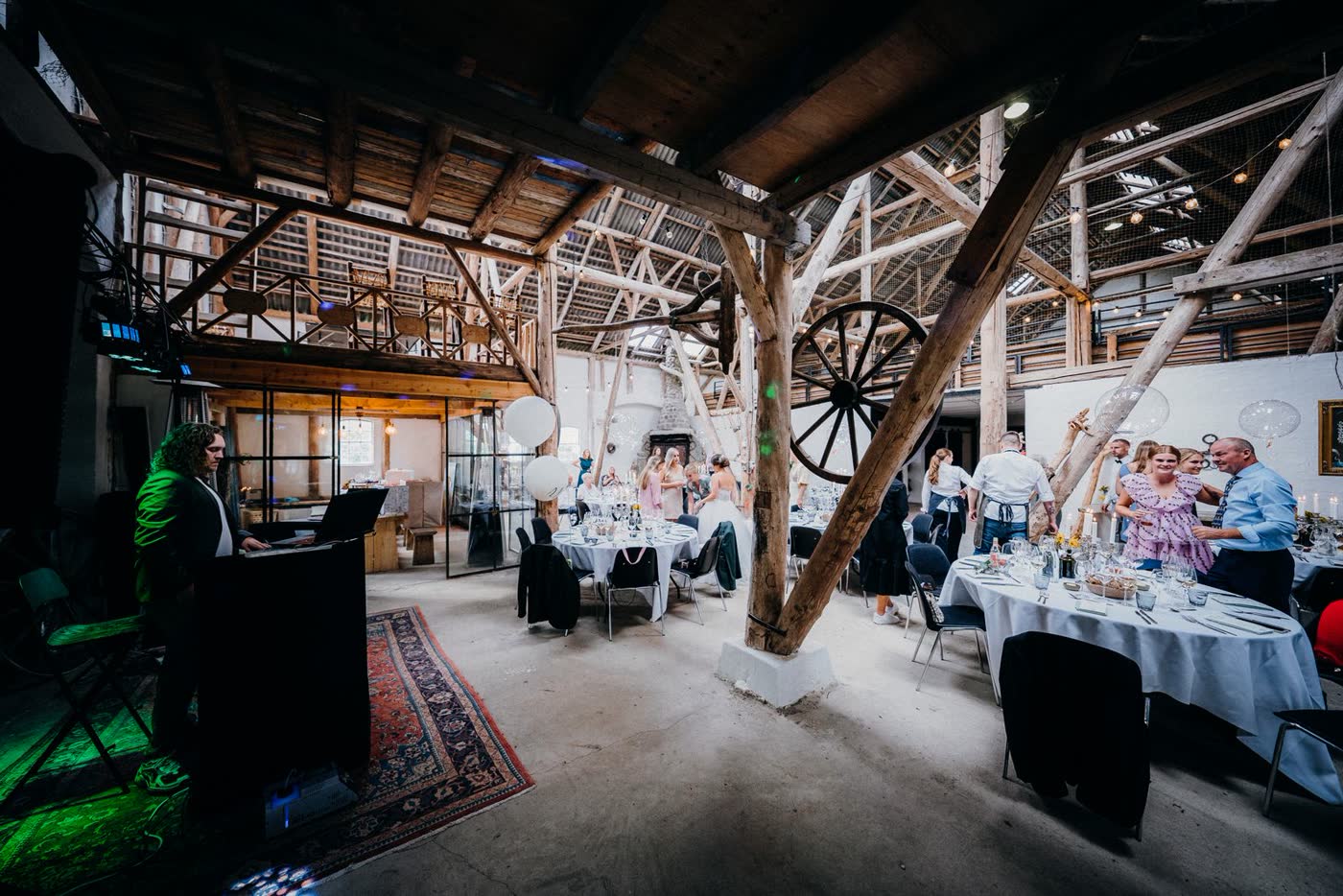 A wedding DJ set up beside the dance floor at a rustic barn reception, with guests and laid tables in the background